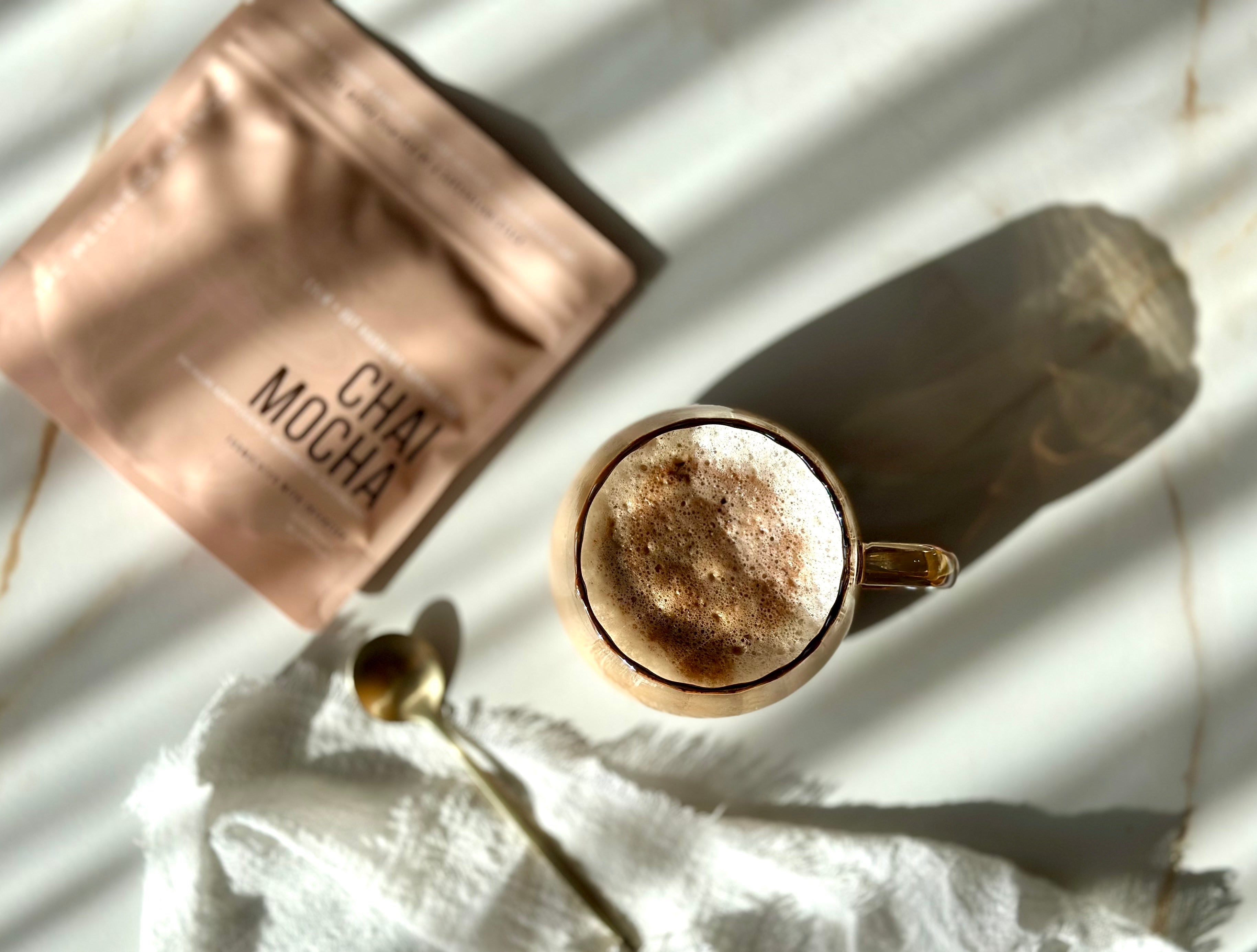 Chai Mocha · Ritual lifestyle — frothy chai mocha in a glass mug beside the pouch, golden hour shadows.